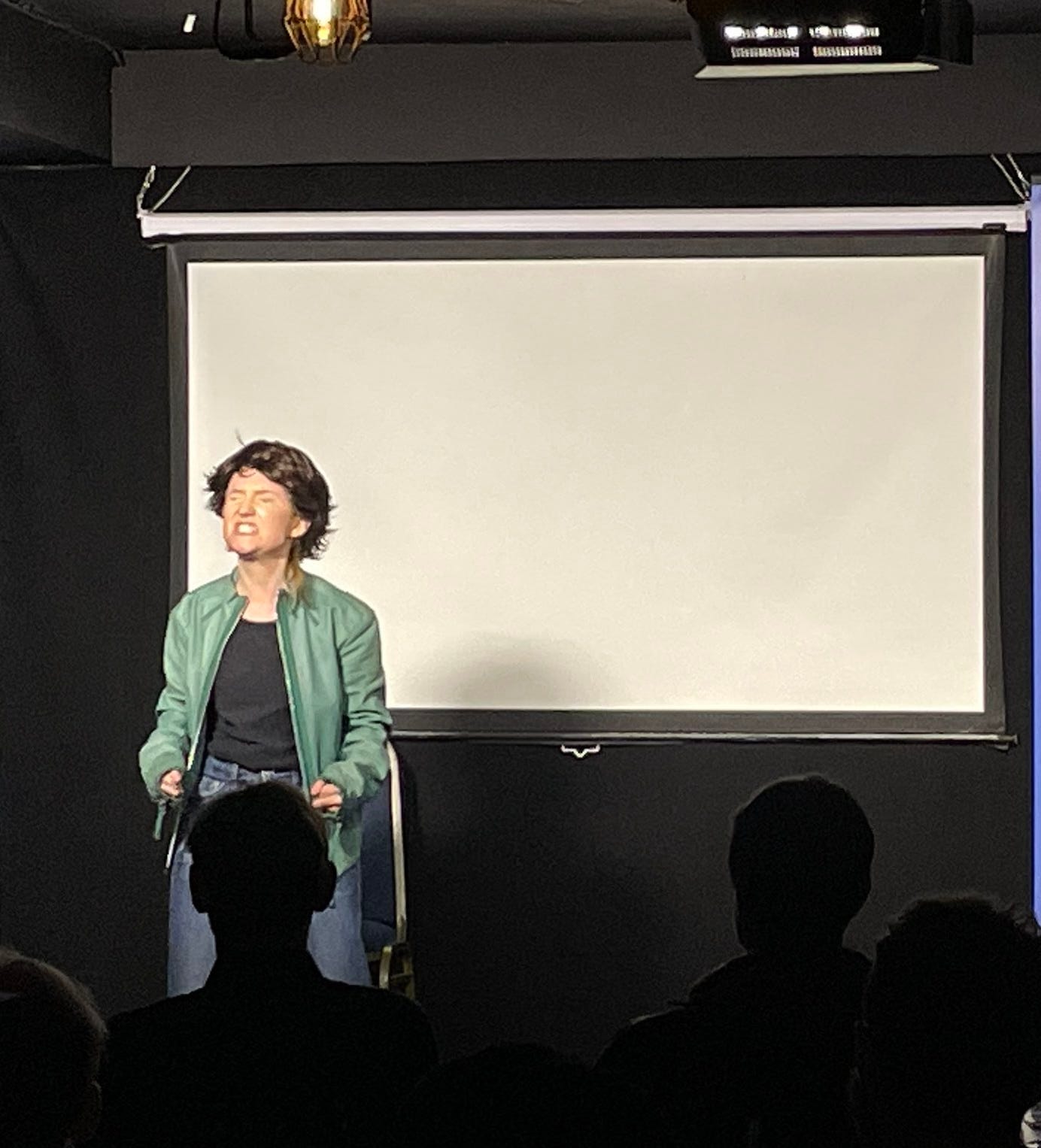 Comedian in a short brown wig and green jacket pulling a strained expression on stage in front of a projector sceen