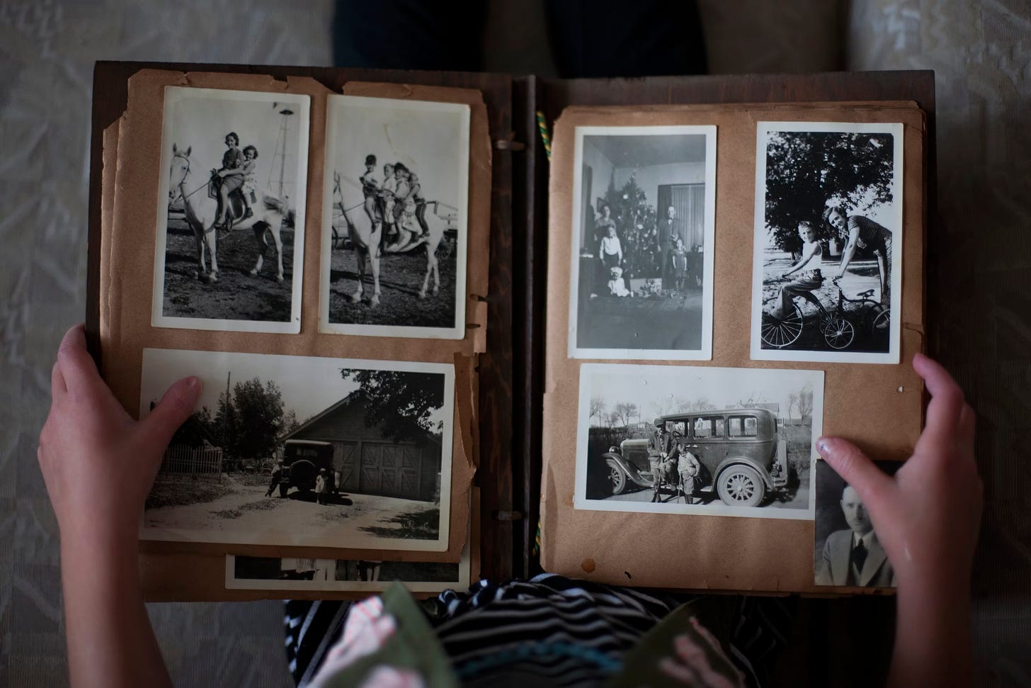 A picture taken from above showing an old photo album held in a child's hands. The album's pages are brown with age and have some stains and tearing around the edges. The photos featured show people on horses and and old-fashioned bicycles and also next to an odl-fashioned car, probably made in the 1930s. One also features a family around a Christmas tree.