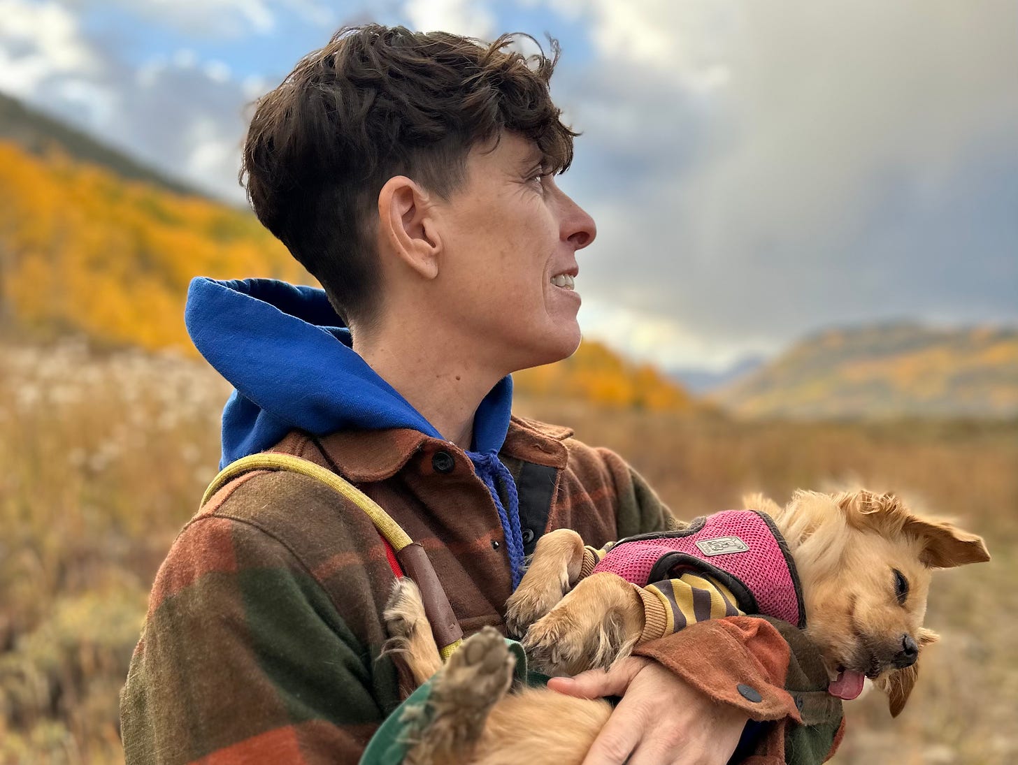 Andrea Gibson is smiling and looking away from the camera while holding their dog Winnie. They are dressed in a red flannel shirt over a blue hoodie, standing amidst golden aspen trees in the mountains. The warm autumn colors create a vibrant yellow backdrop, enhancing the joyful and serene atmosphere of the scene.