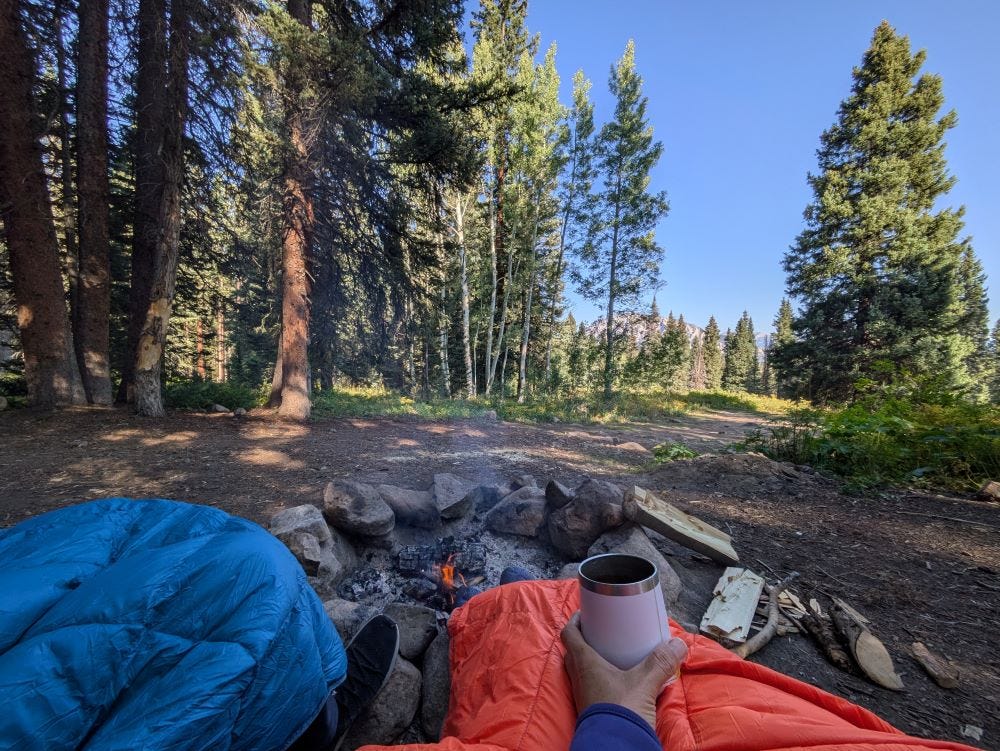 two people sitting by a small campfire in the woods, wrapped in colorful blankets and holding cups of tea and coffee, with a forest and blue sky