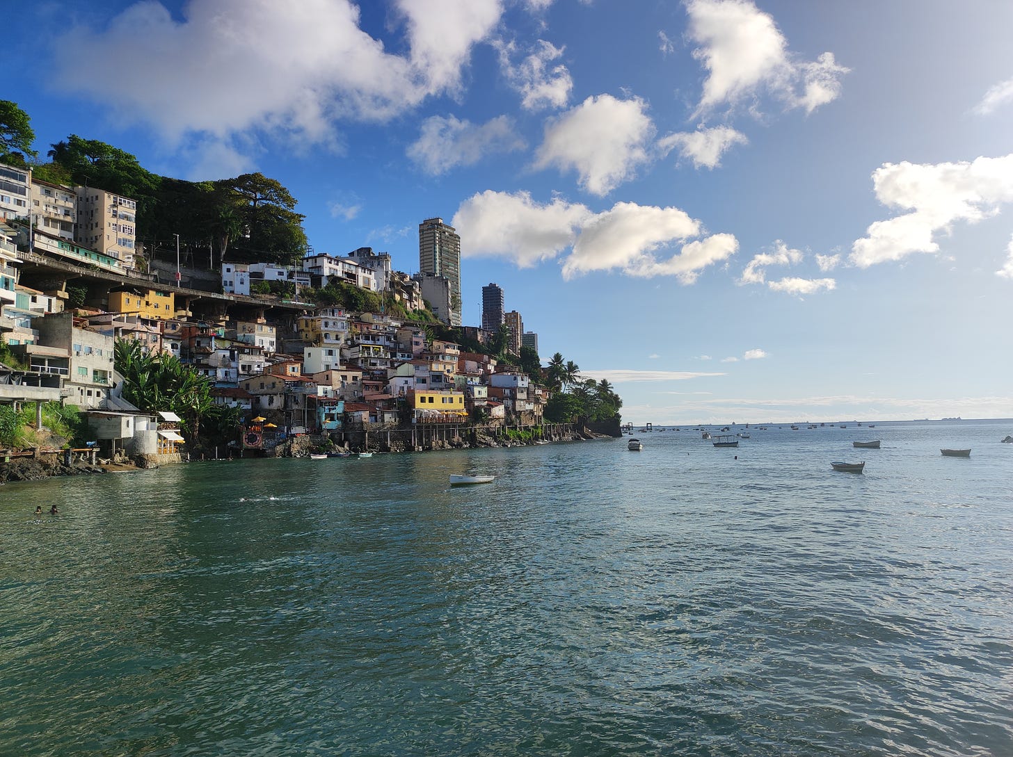 Gamboa, em Salvador, Casas coloridas e humildes a beira mar. Mais distante prédios altos e no mar, azul, alguns barquinhos. É um dia de muito sol;.