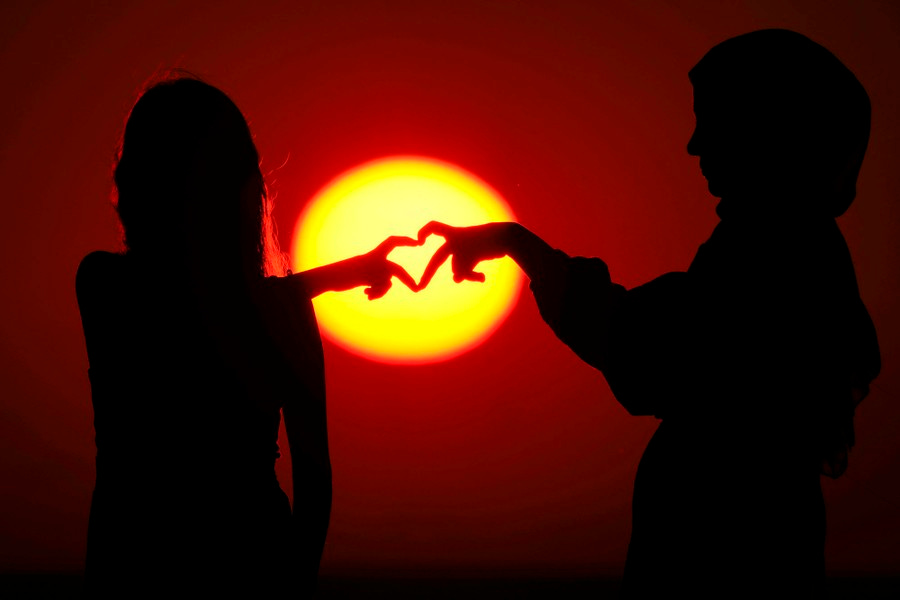 Two young people hold their hands close together, their fingers forming a heart shape in silhouette against the setting sun.