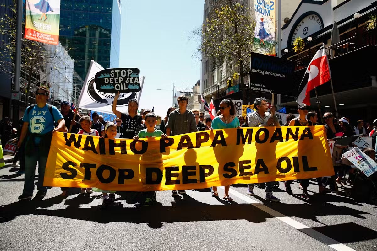 v  photo of people walking down a street protesting. The people at the front are holding an orange banner with the text: Waiho Papa Moana Stop Deep Sea Oil. 