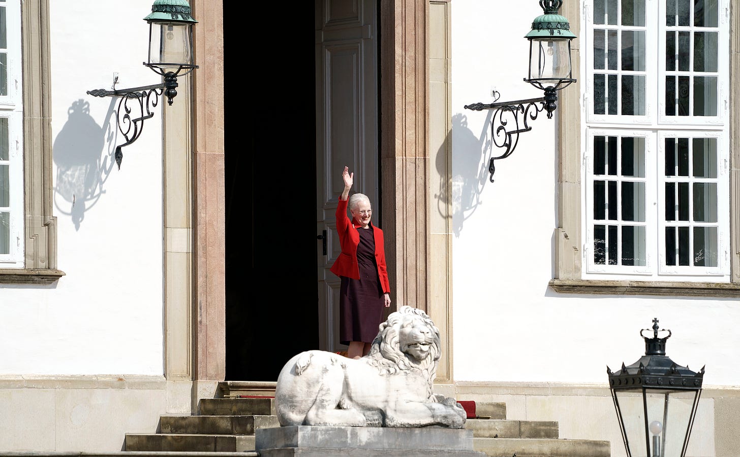 Queen Margrethe waving behind a statue of a lion 