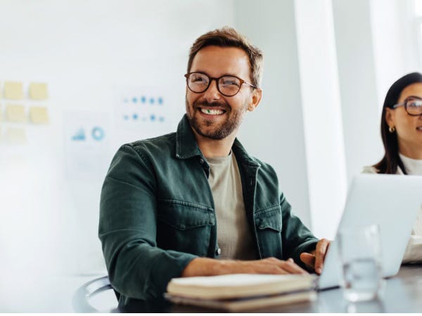Smiling man with glasses at laptop in bright office, with visible spinach stuck between his teeth.