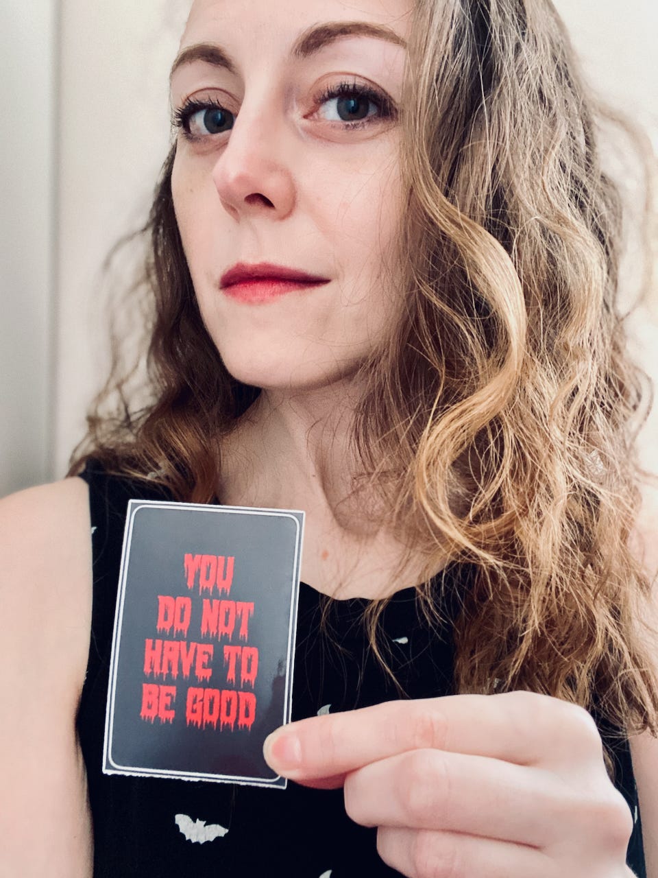 A person holding a sticker and smirking at the camera. The person is pale, with dark eyes and long, wavy hair. They are wearing a black shirt with white bats all over it. The sticker is a black rectangle with text in dripping, bloody red letters: YOU DO NOT HAVE TO BE GOOD