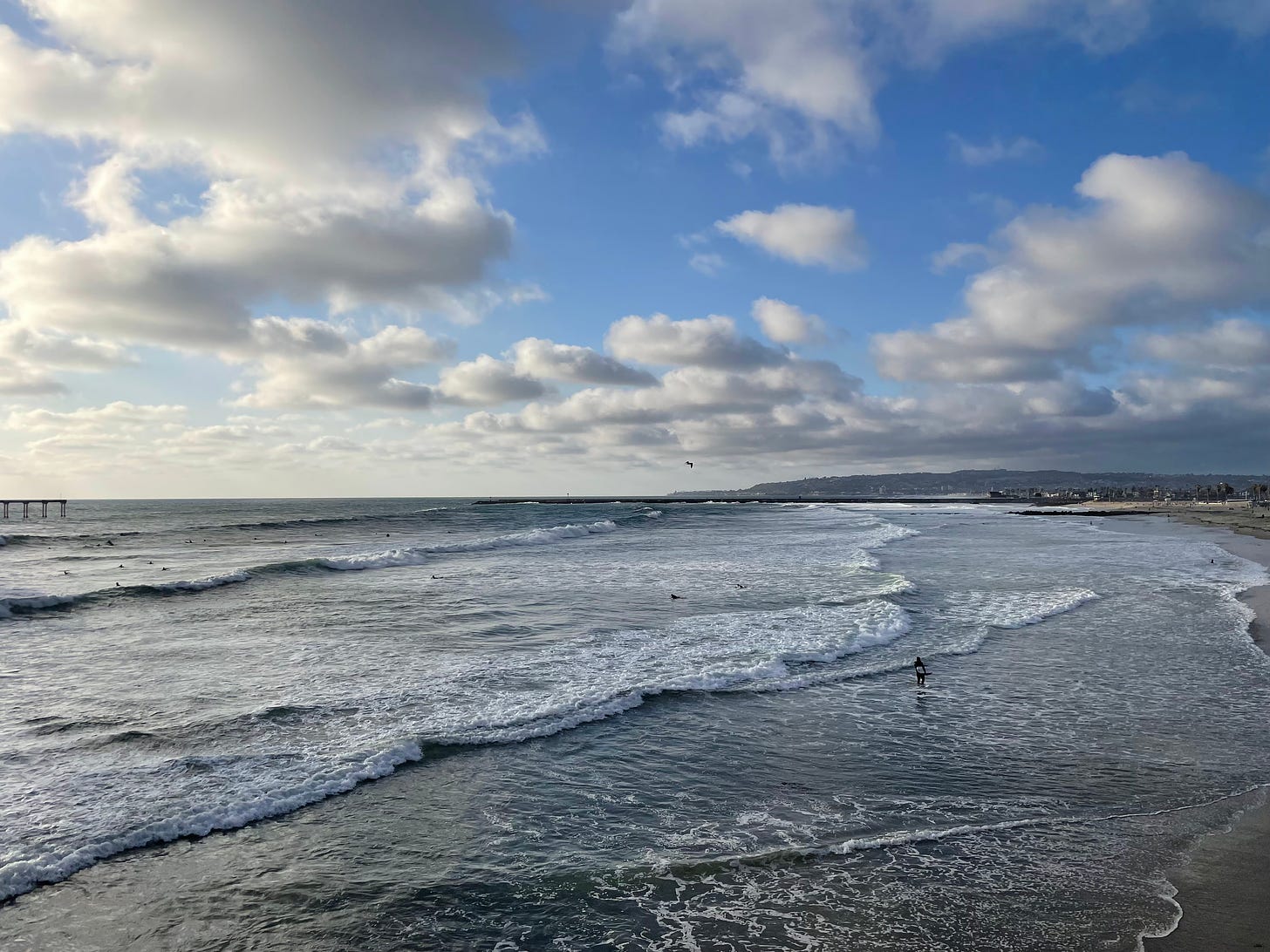 Waves of the ocean shown from above. The sun begins to set, with clouds scattered throughout the blue sky. 