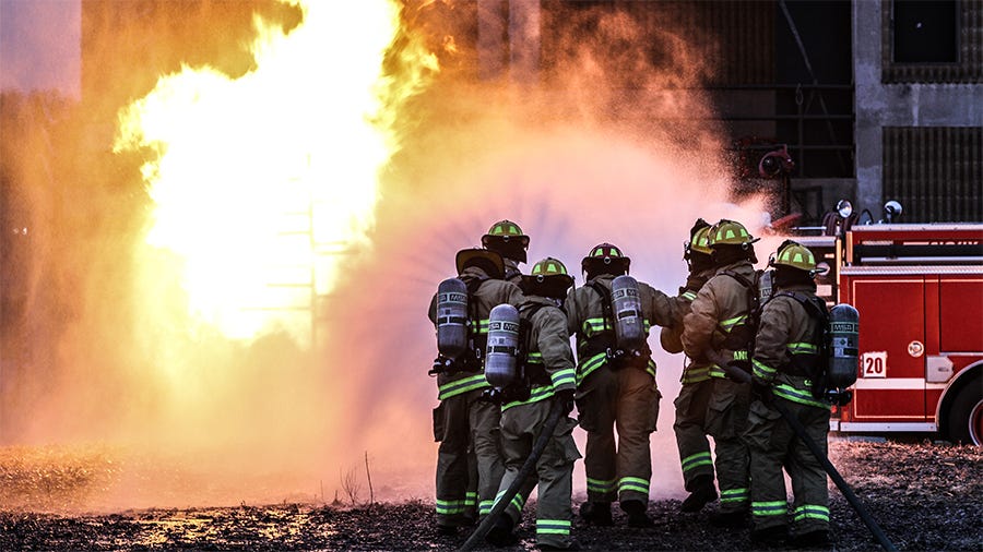 Firefighters in protective gear using a hose to extinguish a large blaze near a building, with a fire truck parked in the background.