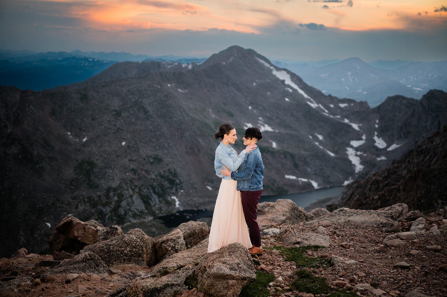 Shohreh and Jessie are wearing jean jackets over their wedding outfits and standing looking at each other with a huge mountain range and beautiful sunset visible behind them