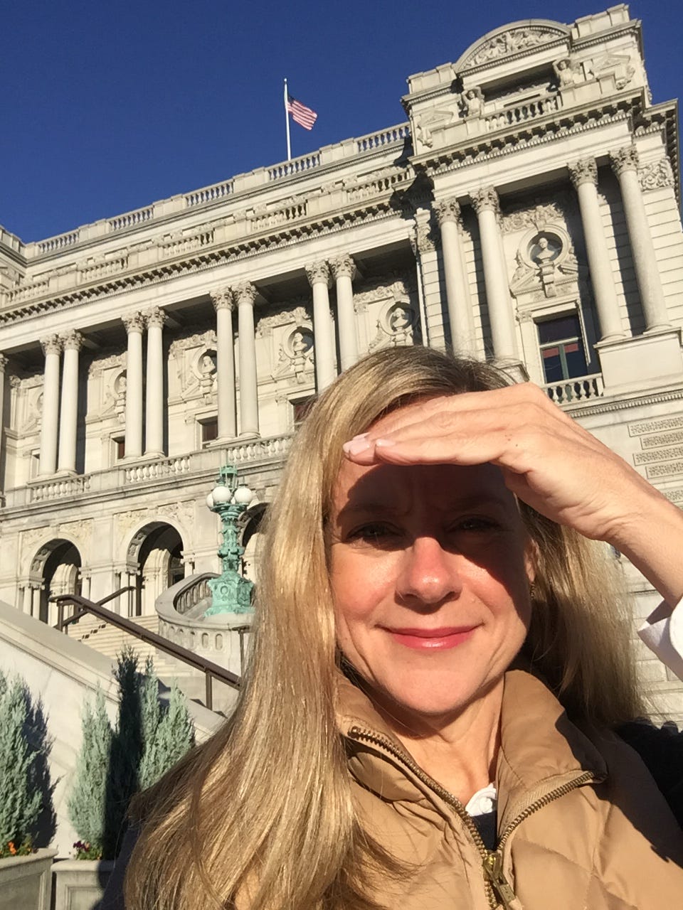 Woman shielding her eyes from the sun, standing in front of the Library of Congress with white marble pillars and an American flag in the backround
