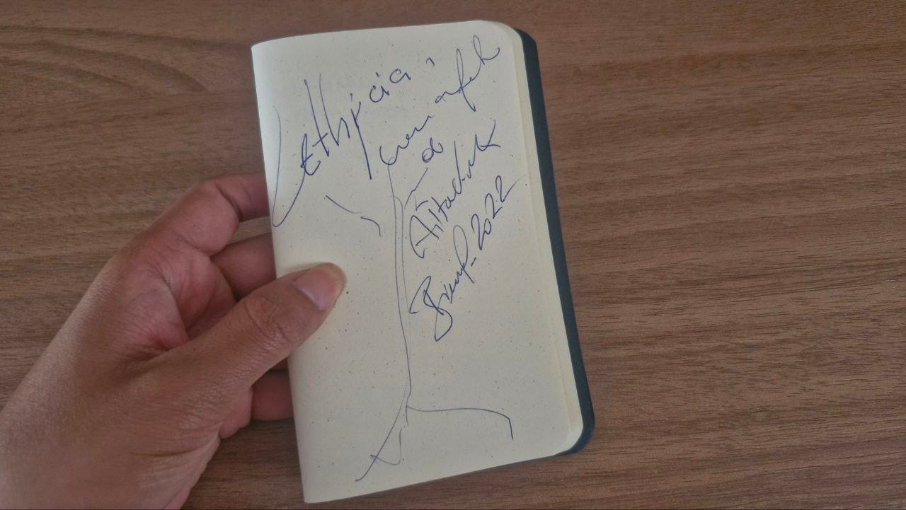 Sobre uma superfície de madeira, minha mão esquerda segura uma pequena caderneta com folha sem pauta, exibindo numa página, um autógrafo endereçado a mim, Lethycia, escrito ao lado do desenho de uma árvore. Sobre uma superfície de madeira, minha mão esquerda segura uma pequena caderneta com folha sem pauta, exibindo numa página, um autógrafo endereçado a mim, Lethycia, escrito ao lado do desenho de uma árvore.