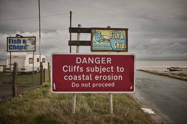 A red sign along a road reads "DANGER: Cliffs subject to coastal erosion. Do not proceed." Another sign above it advertises fish and chips. 
