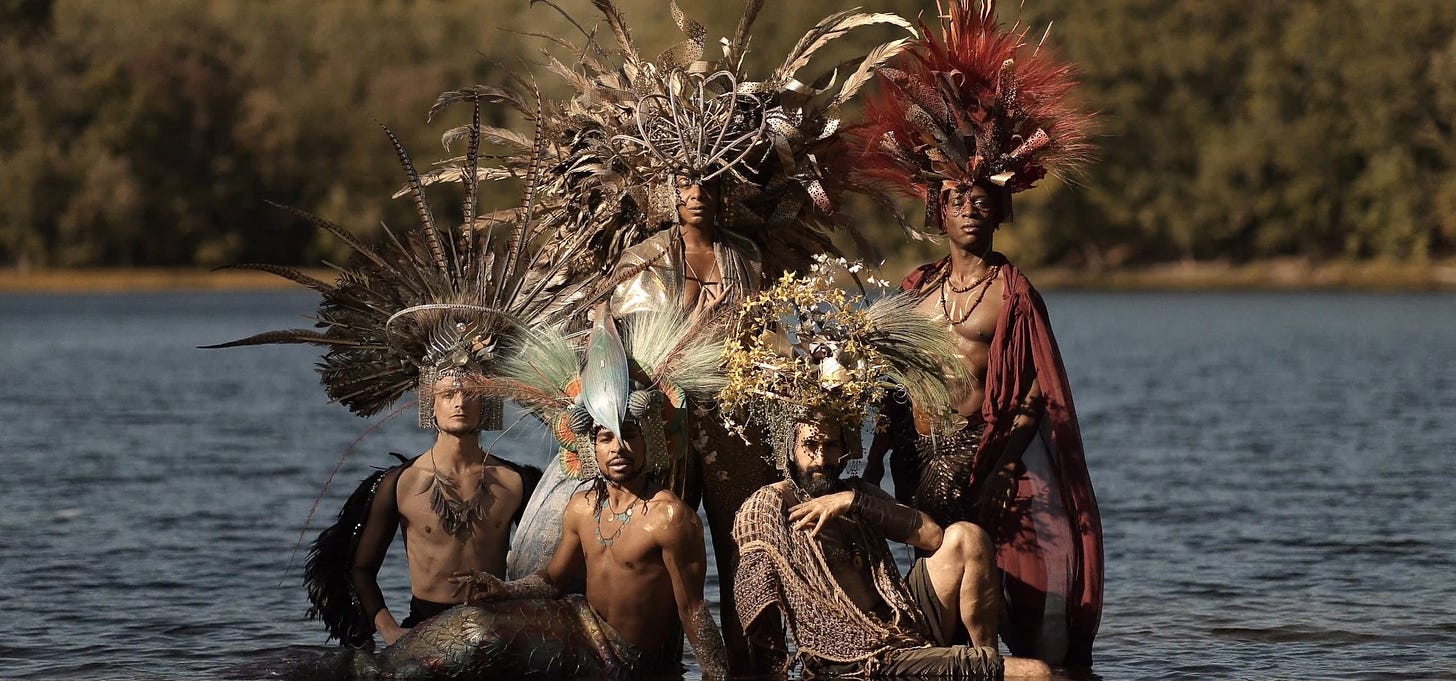 5 performers are in the shallows of water with trees in the blurry background. They wear ornate headdresses of flowers and feathers, looking serenely at the camera.