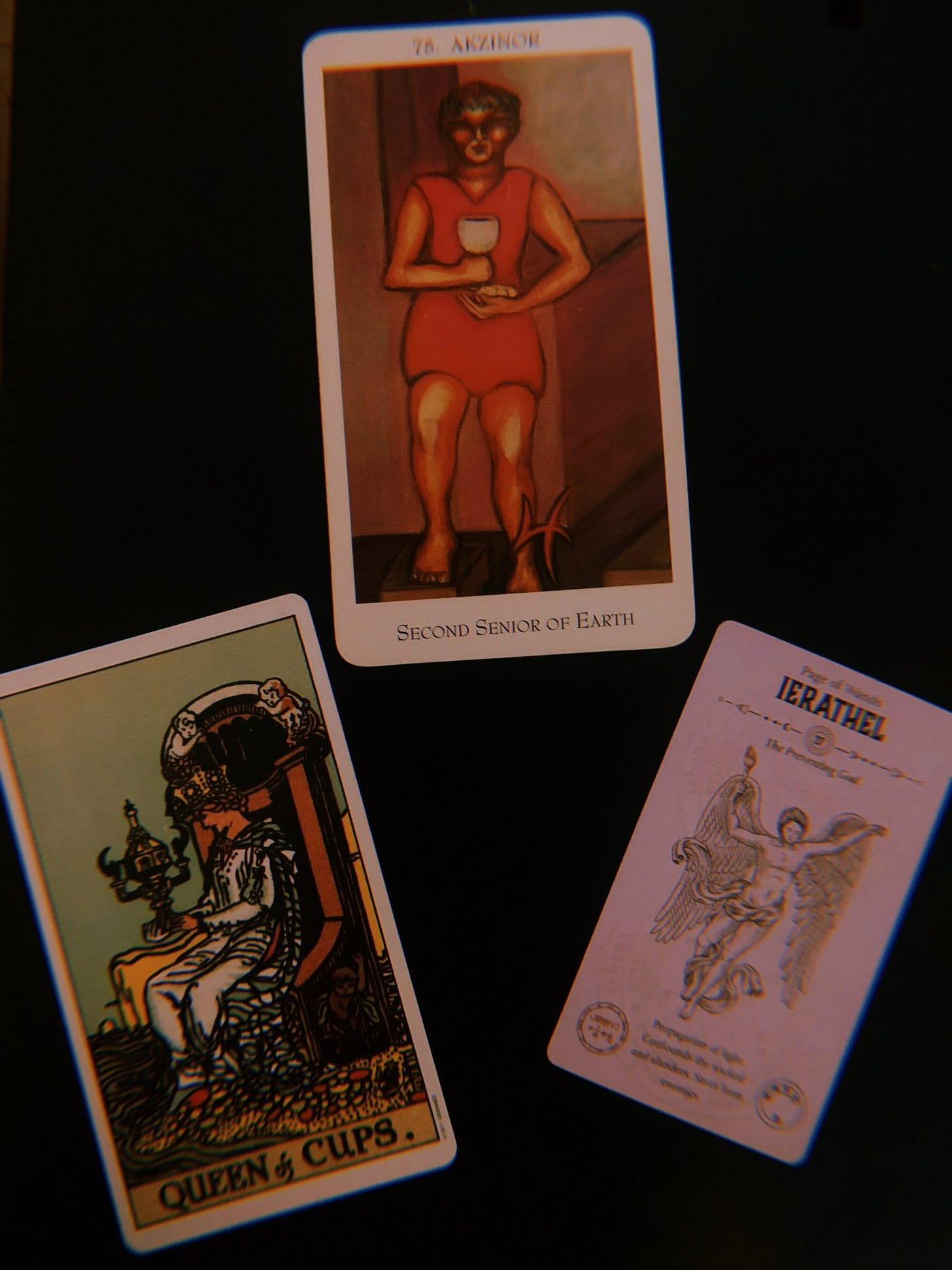 Three difference divinatory cards sit on a black background. On the left a Rider-Waite tarot card of the Queer of Cups. The queen sits on her throne decorated in sea nymphs on the beach, just far enough that the water does not touch her feet. She stares with contempation at an ornate cup decorated with angels. The middle card is the Enochian card, 76. ARZINOR Second Senior of Earth. A tall, masculine figure wears a short pink dress and stares forward with concentration. He holds a yellow goblet and wears a laurel on his head. On the right is an Abrahamic angel card of IERATHEL the Preventing God. The toned angel is a white, masculine figure with hands aloft, one holds a torch. They wear only a flowing sash around the hips and has outstretched bird wings. 