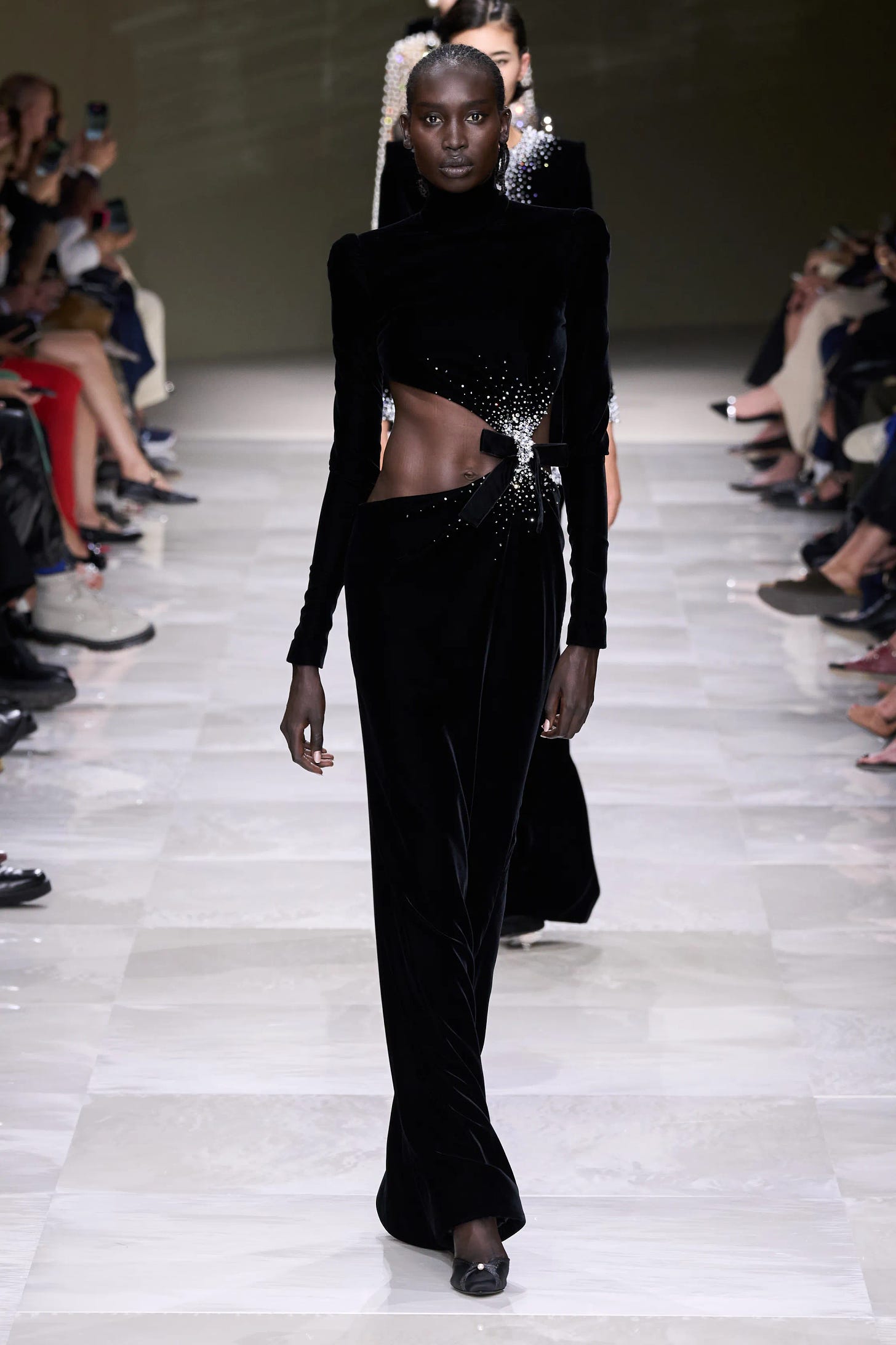 A slender dark-skinned model walks down a pale gray runway wearing a black velvet dress with slender sleeves and an exposed midriff above a floor-length skirt.