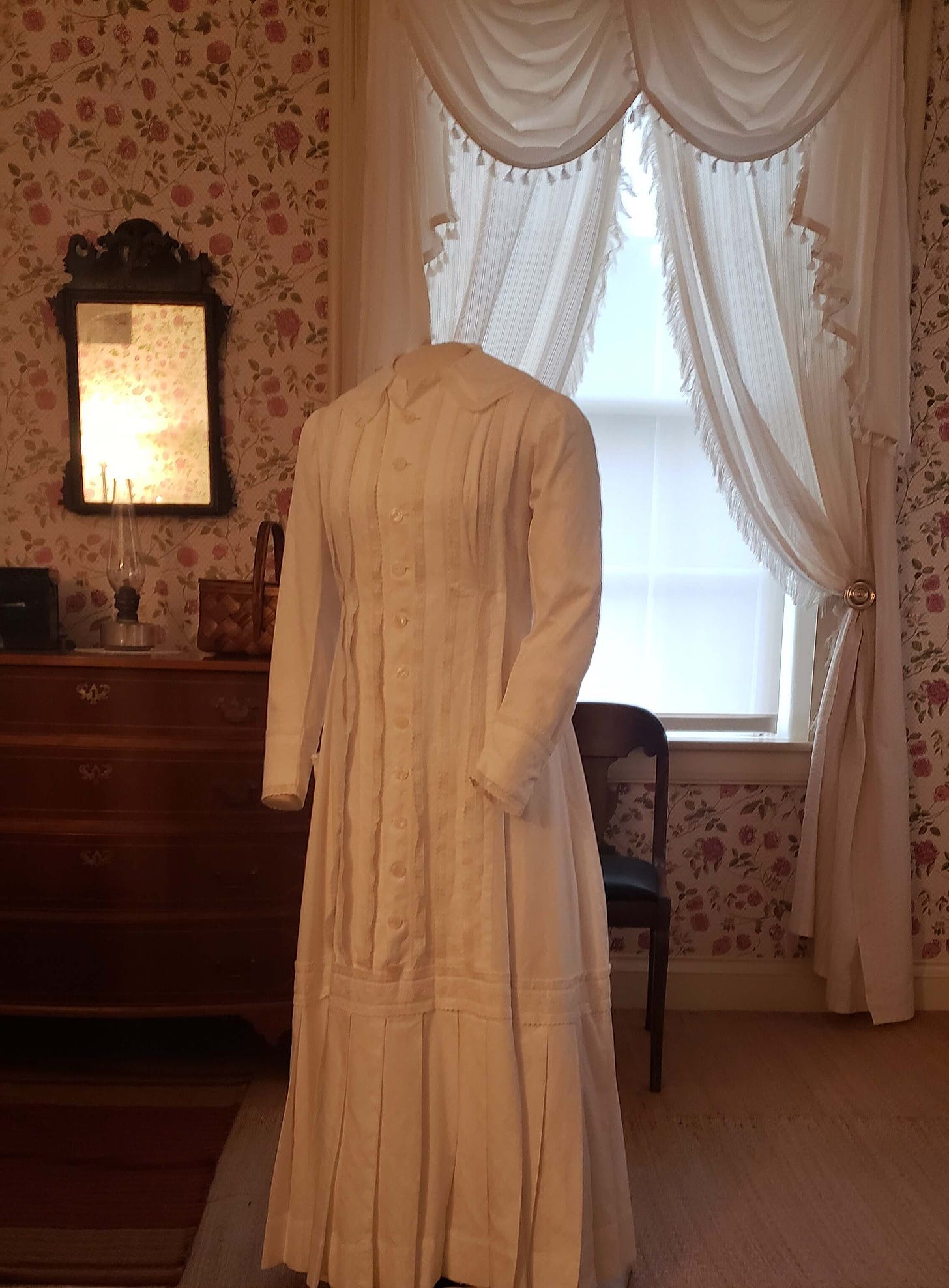 A small form with a replica of Emily Dickinon's white dress. Long-sleeved, with a Peter Pan collar and pleats all the way down to the knees, then a pleated skirt. Buttons all the way down to the skirt. Behind, her dresser and mirror, and a window with beige curtains. The wallpaper is packed with deep pink roses. 