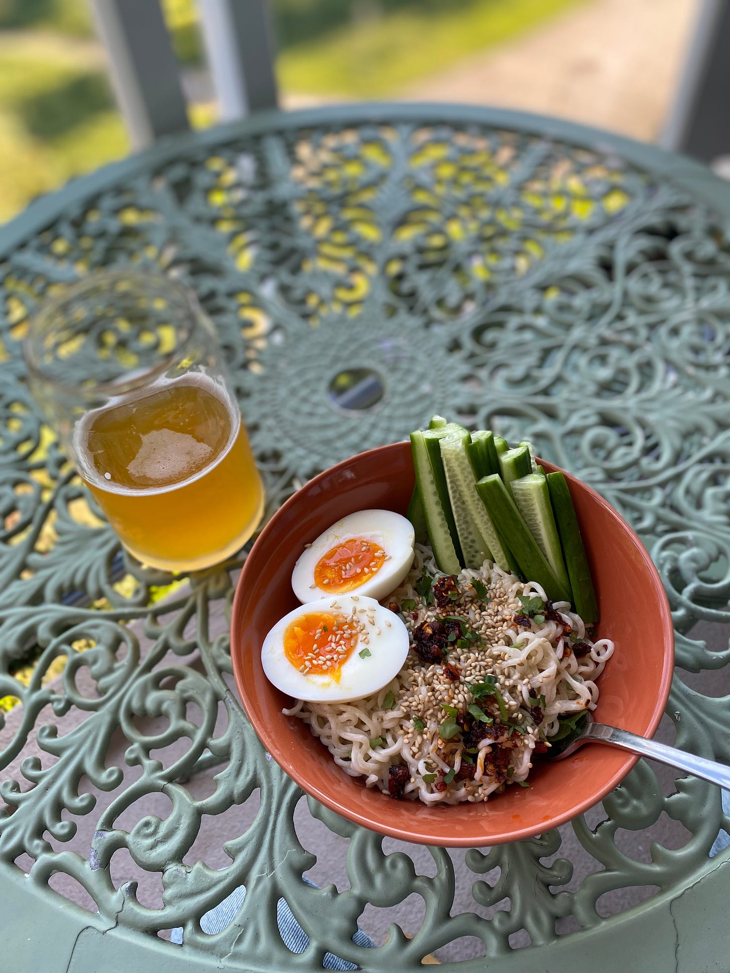 An orange bowl of ramen noodles in sauce with chili crisp, sesame seeds, and cilantro on top. Arranged around the sides are cucumber sticks and two halves of a boiled with an orange, jammy yolk. Next to it on an outdoor table is a glass of beer.