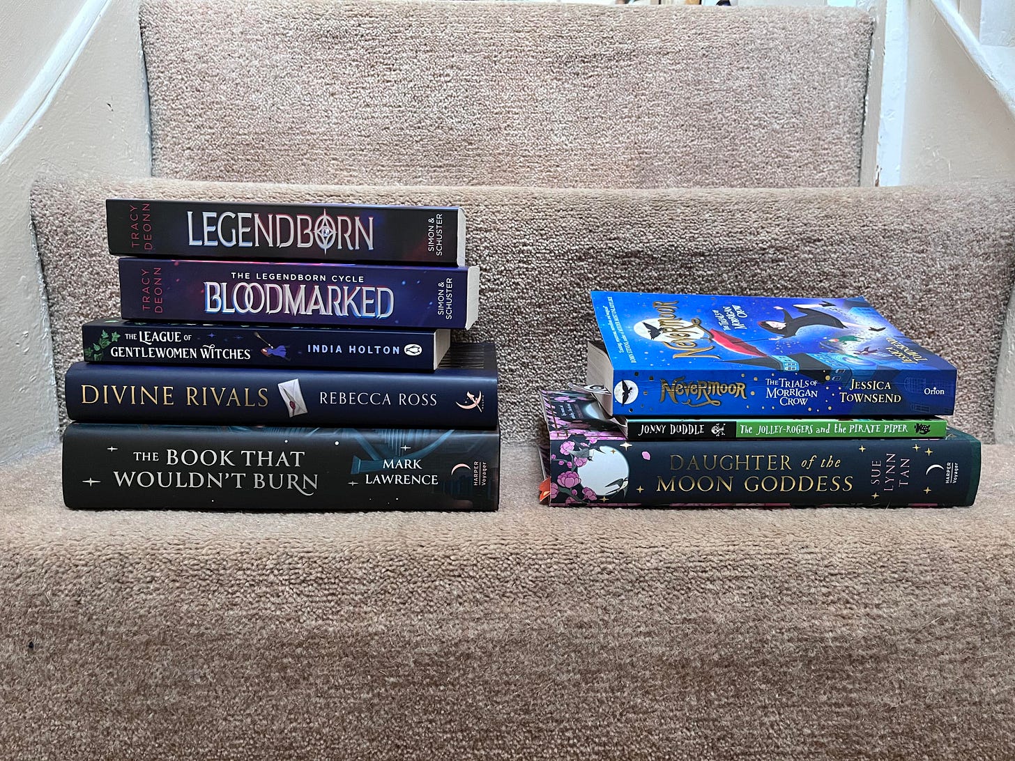 Two piles of books sitting next to each other on cream coloured carpet. The pile on the left includes The Book That Wouldn't Burn; Divine Rivals; The League of Gentlewomen Witches; Blookmarked; Legendborn. The pile on the right includes: Nevermoor; The Jolly Rogers and the Pirate Piper; Daughter of the Moon Goddess.