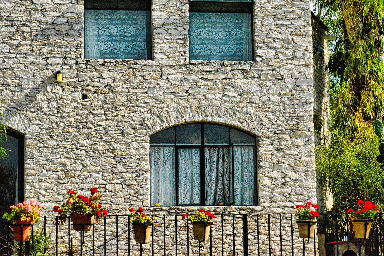 a stone house with two windows on the second floor and one on the first floor with lace windows and an iron fence in front with pots of red geraniums
