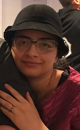 Picture of Emmalia Harrington. She is wearing a black hat and burgundy shirt and affectionately holding the arm of a second person, who is out of the frame.