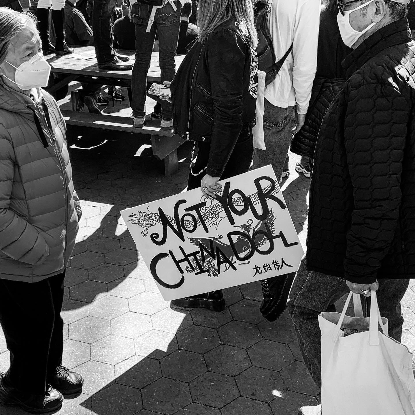 a black and white photo of people walking in a crowd, one woman is holding a sign that says "Not Your China Doll."