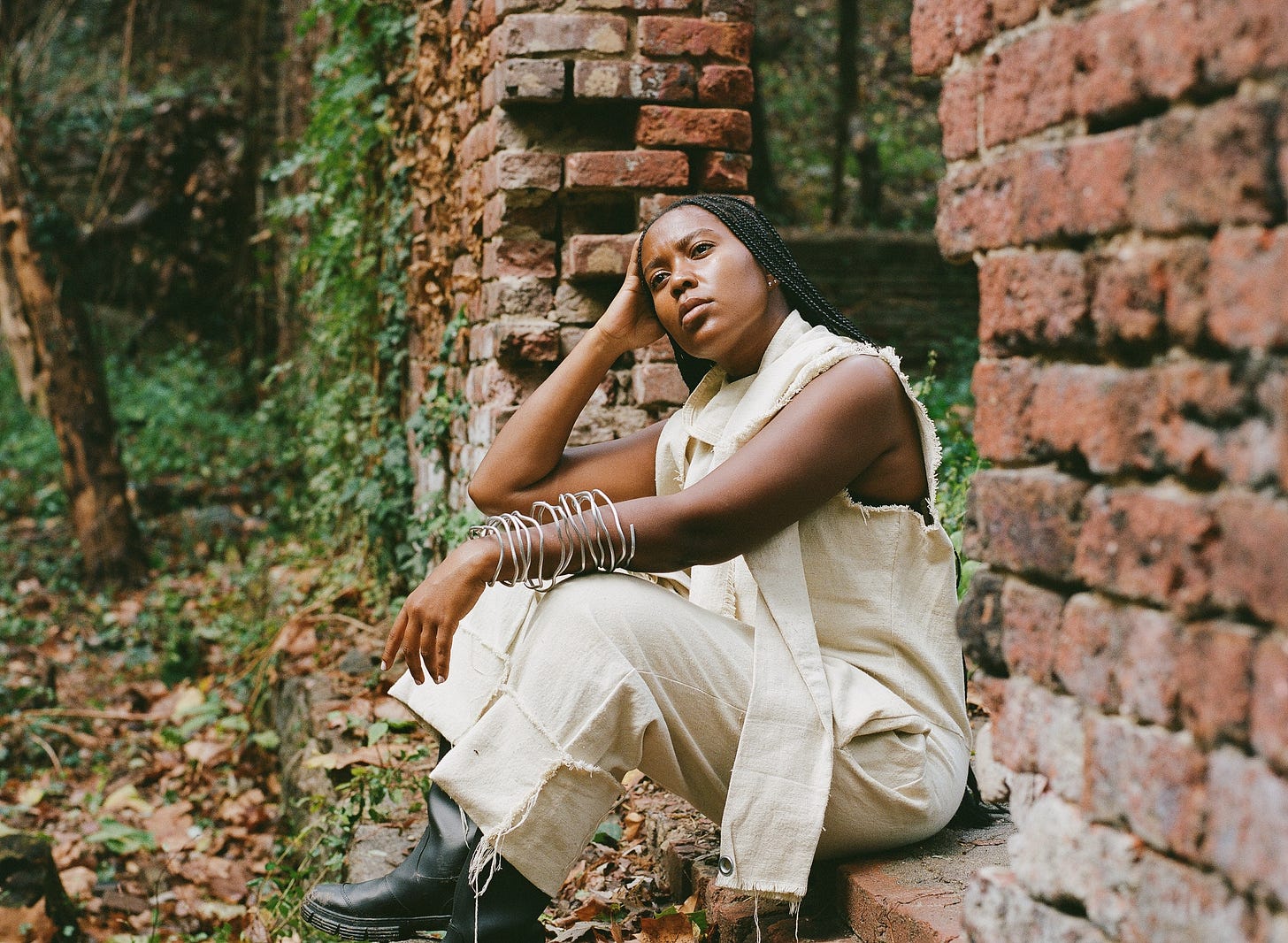 Image Description: I am wearing long black braids, an unbleached cotton jumpsuit canvas jumpsuit I made from drop cloth, black rubber boots and an armature wire arm cuff sculpted around my arm while gazing away from the camera and sitting inside an opening of a decaying brick wall being reclaimed by the forest. Photography: Chukwudumebi Ezefili