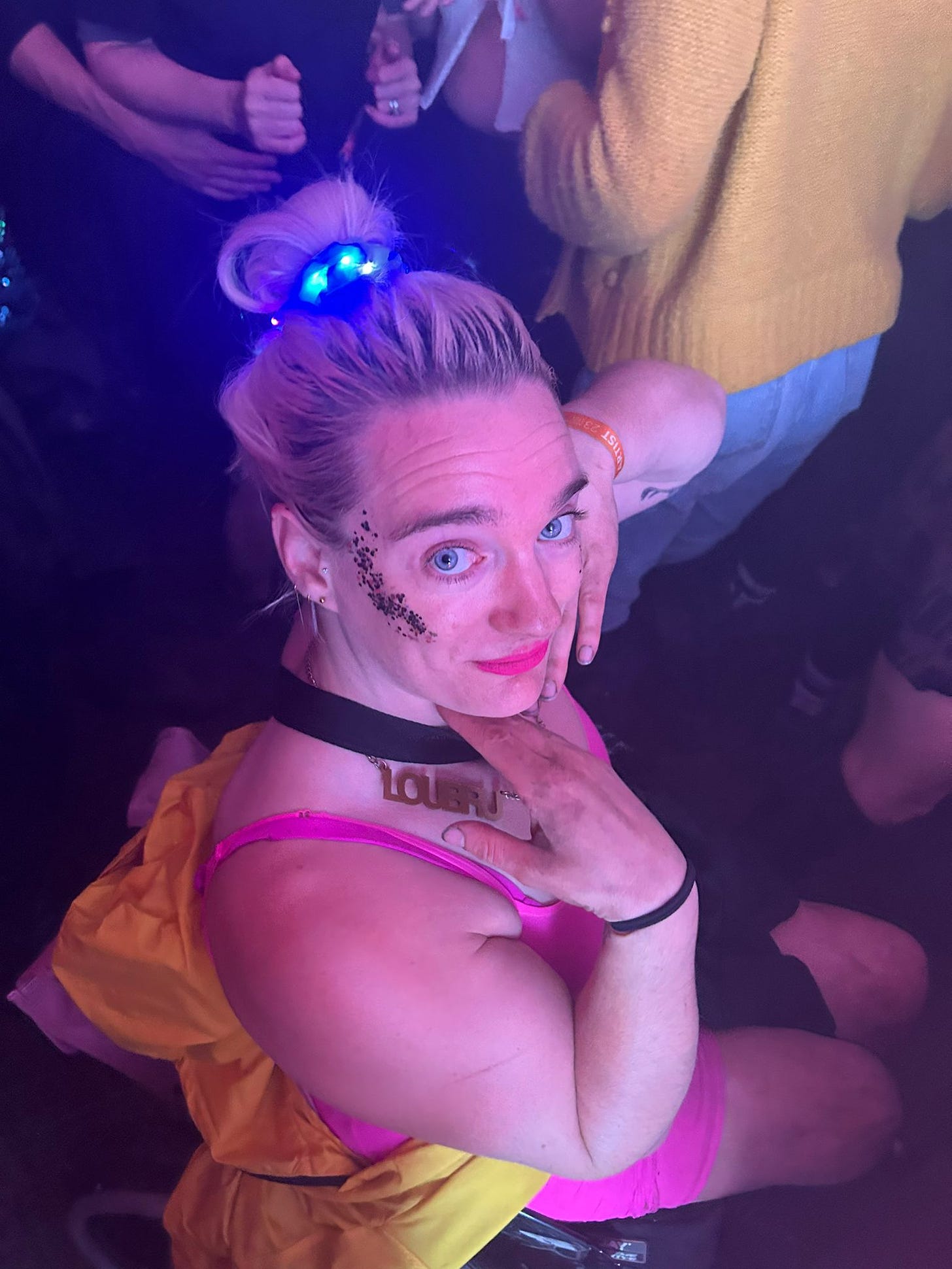 I am wearing a pink bodycon dress and posing with my hands under my chin. Covered in mud and glitter, I wear an LED scrunchie in my hair and have a rain coat tied around my waist as I sit in the middle of a festival crowd. 