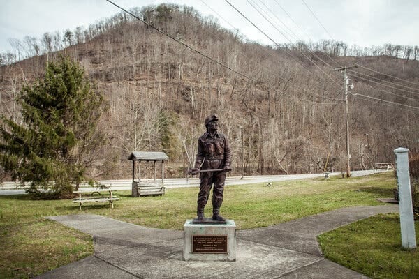 A bronze statue of a coal miner dressed in work clothes, helmet and head lamp and holding a pick ax is at the center of a green space. 