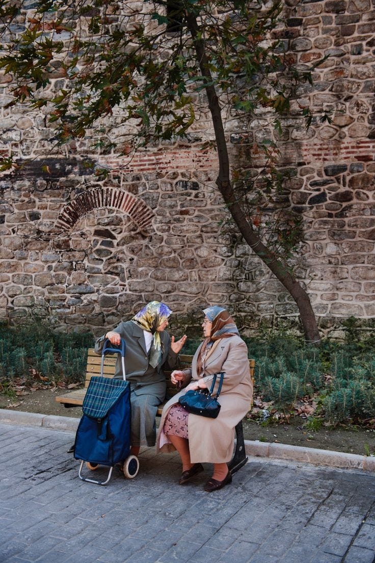This may contain: two women sitting on a bench next to each other near a tree and brick wall