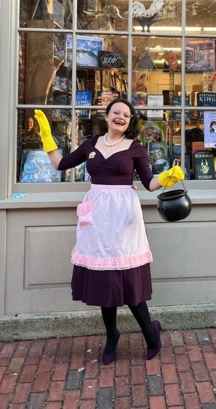 A white woman in a purple dress with a pink apron stands in front of a bookstore. The woman is wearing yellow kitchen gloves, in one hand she holds a plastic halloween cauldron.