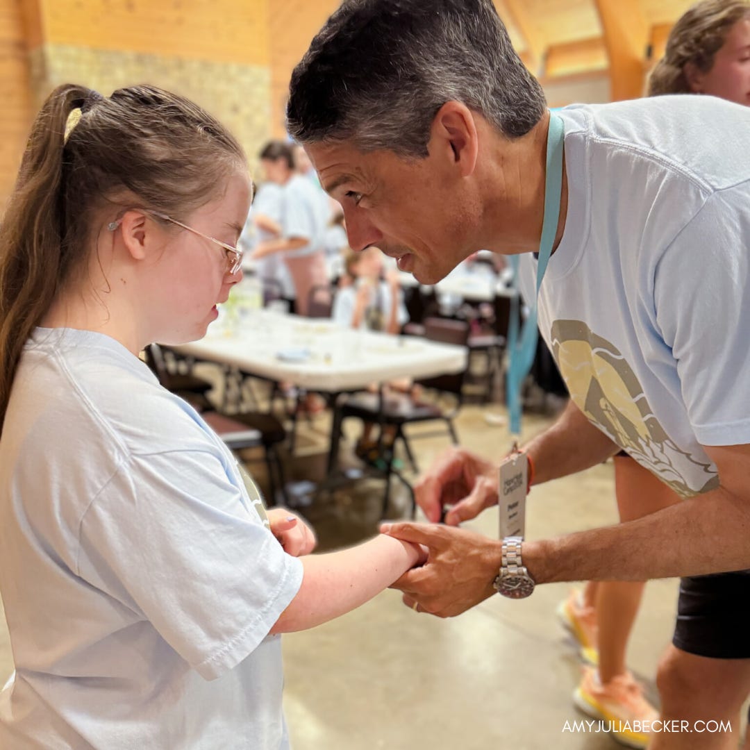 Peter is gently holding Penny’s hands as he makes the shape of a cross on her hands. He is leaning in slightly. Both are wearing light blue Hope Heals T-shirts. In the background, other people can be seen in a room with tables, creating a warm, communal atmosphere. The mood of the scene is caring and supportive.