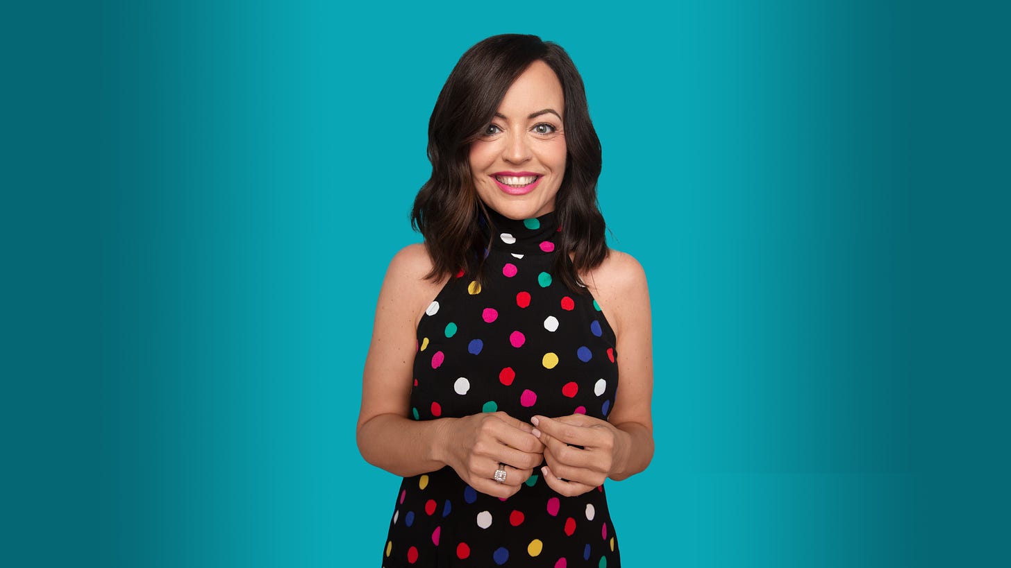 A photo of me wearing a black halter top dress with multi-colored polka dots. My hair is shoulder length and dark and I am smiling wide.