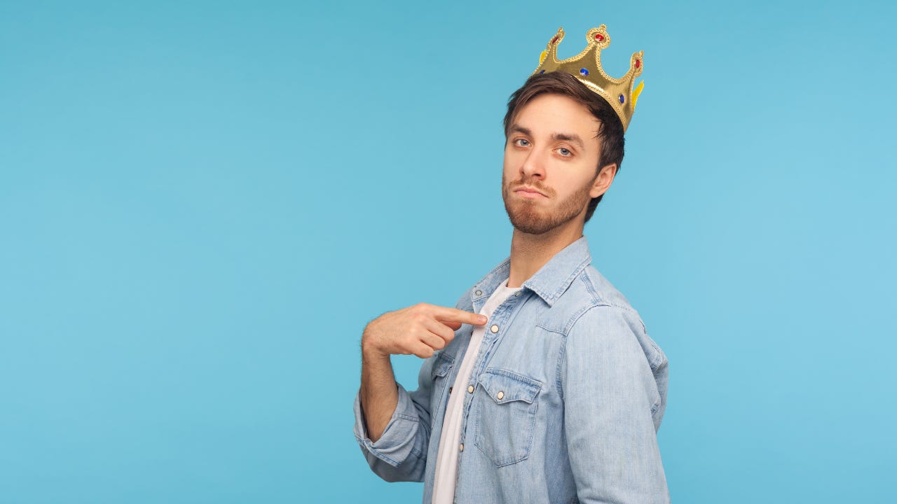 An arrogant man wearing a crown and pointing at himself.