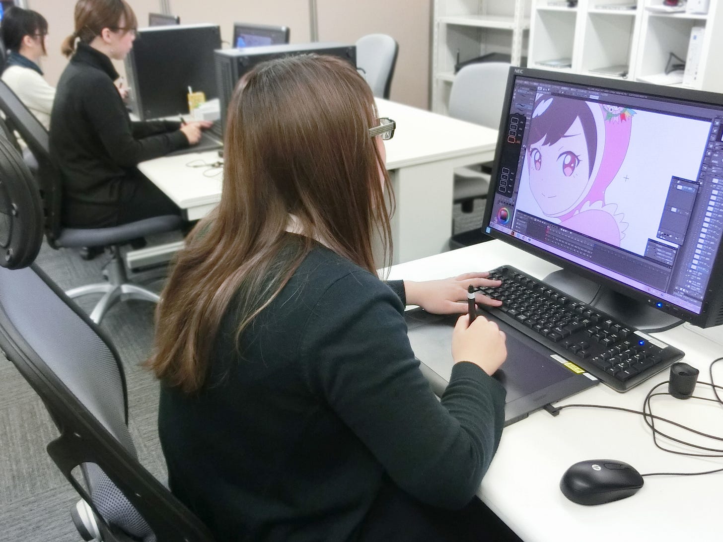 A female animator in the foreground sits facing away from the camera at a computer. She is drawing on a digital pad with a stylus in her right hand, and a color drawing is displayed on the monitor screen. Her left hand is hovering on a keyboard.