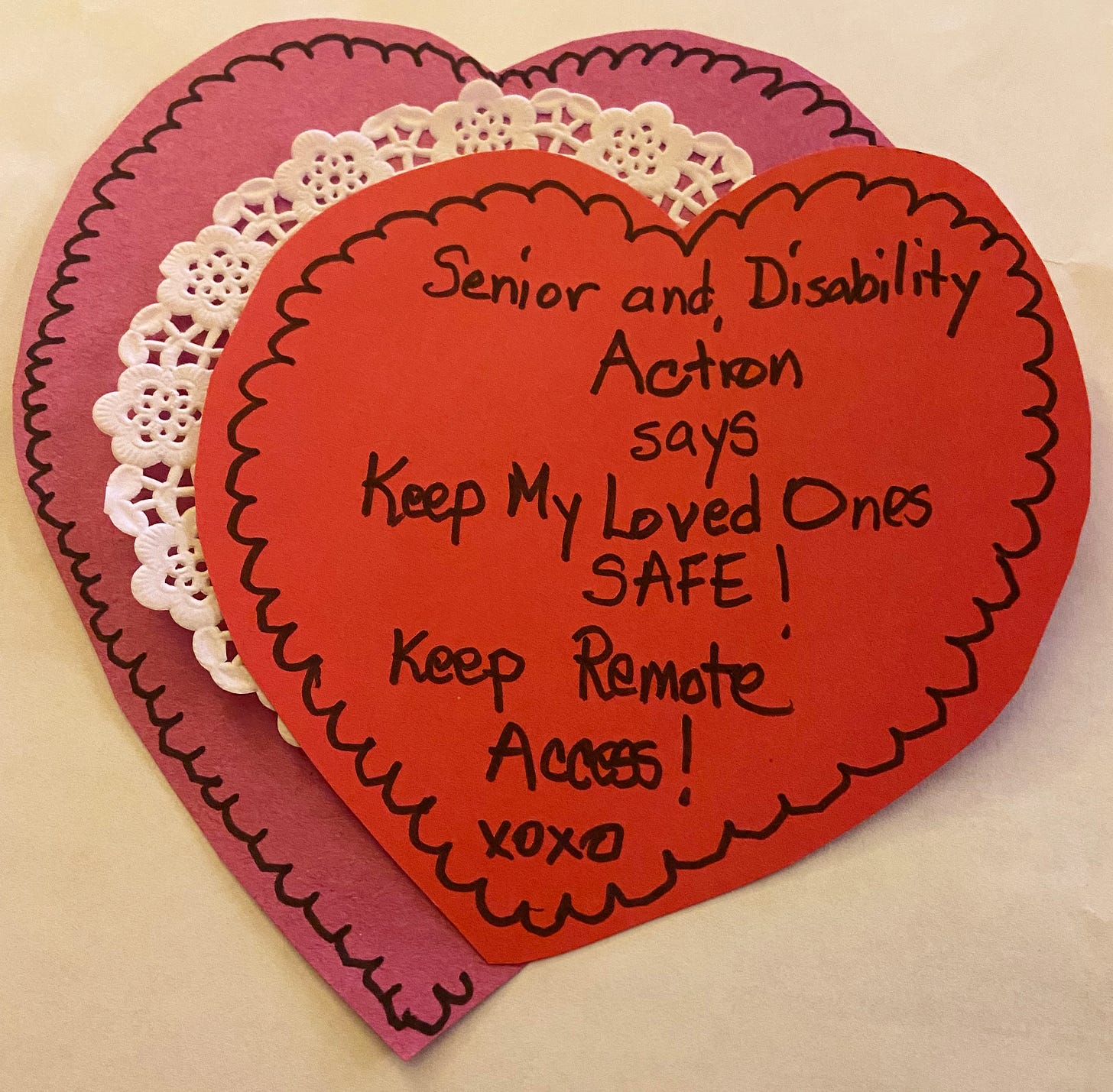 The photo is of a homemade Valentine card, a double construction paper heart sandwiching a doily, decorated with black marker and the words Senior and Disability Action says Keep my loved ones safe! Keep Remote Access! xoxo