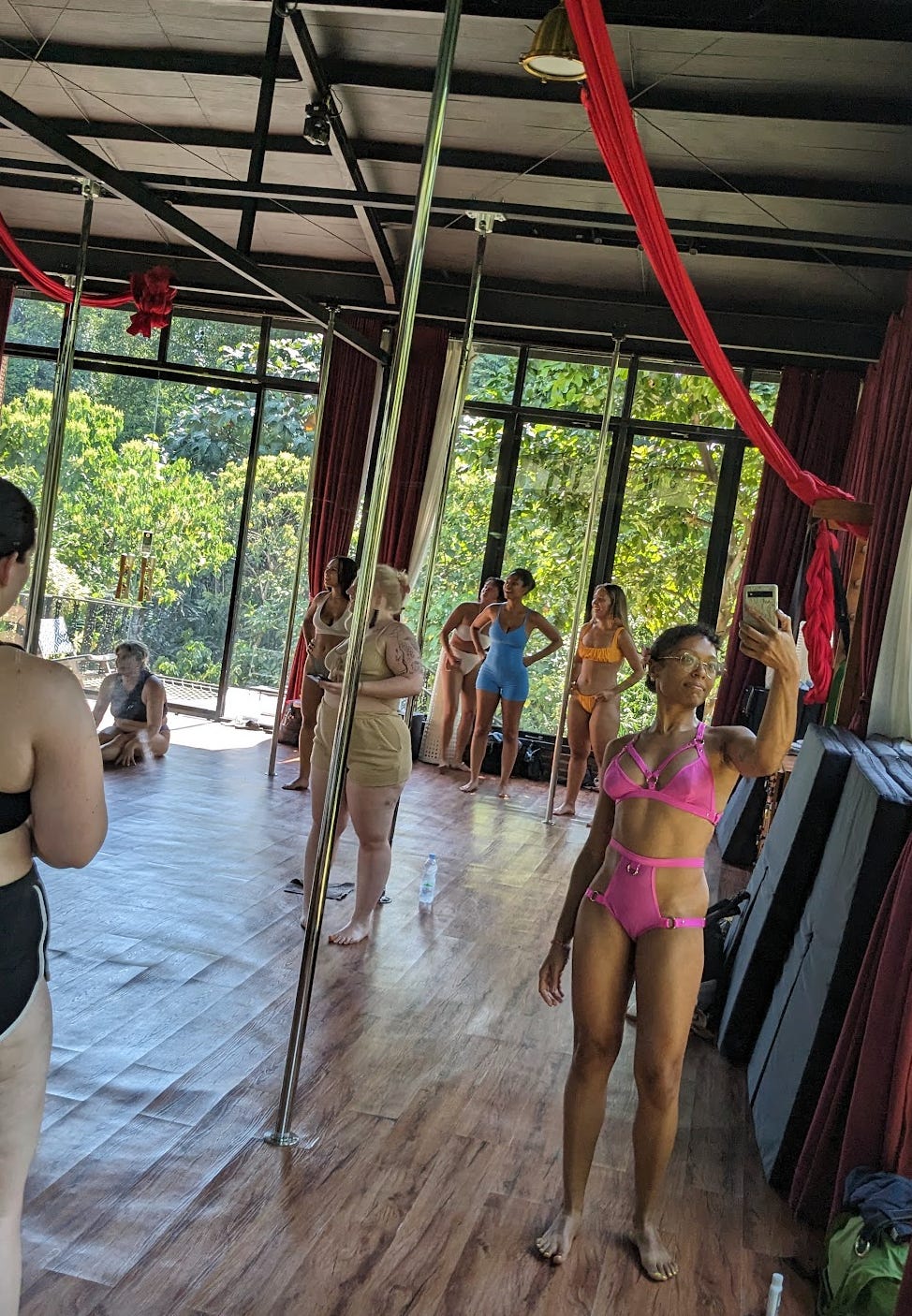 Nathalie snaps a photo of herself in front of the large window in sun-filled pole dancing studio that is filled with other femmes. Nathalie is wearing a bright hot pink underwear set that has silver buckles. Some people in the background are wearing their own colorful sets including a sky blue sports bra and biker shorts and mustard yellow underwear set