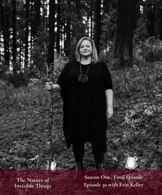 Erin is standing in the woods between two lanterns. She is wearing a black tunic and a black leather tassel necklace. In her right hand she holds her forked stang. 