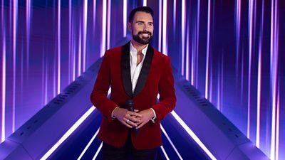 A man wearing a velvet red suit jacket over a white shirt stands on a futuristic stage holding a microphone, against a backdrop of purple and white beams of light 