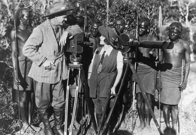 A vintage photograph of a European couple dressed in hats look at one another as they pose with two cameras in front of a row of shirtless African men holding spears, some of them bemused.