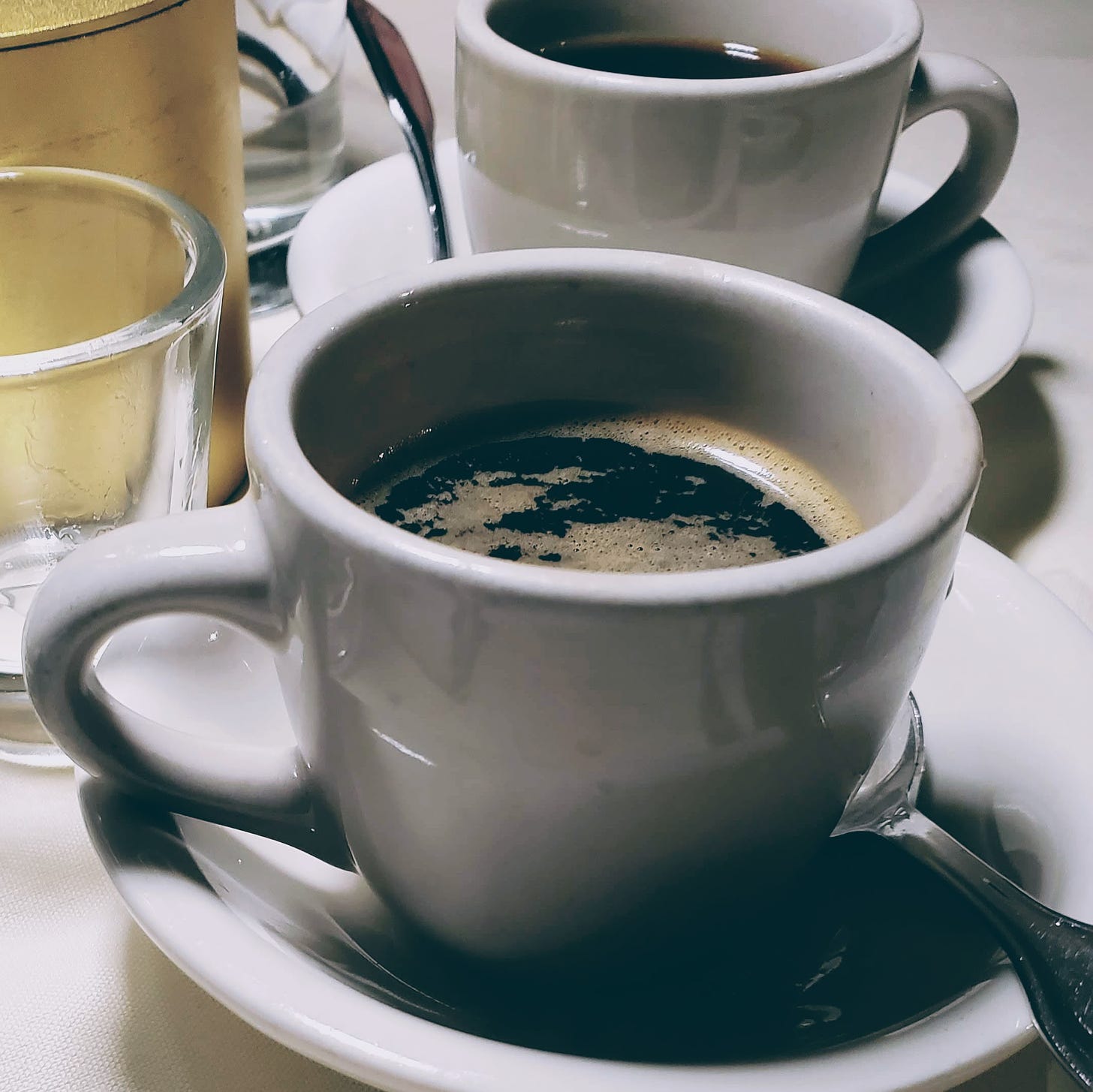 two white cups of coffee, sitting on saucers, with spoons, and glasses and a white table cloth in the background