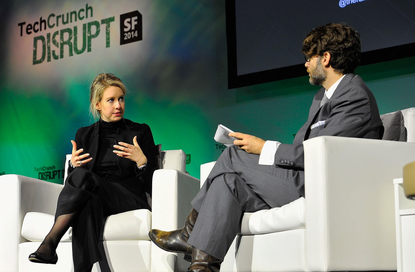Elizabeth Holmes, wearing an all-black suit, sits on a white armchair on the TechCrunch Disrupt stage in San Francisco. 