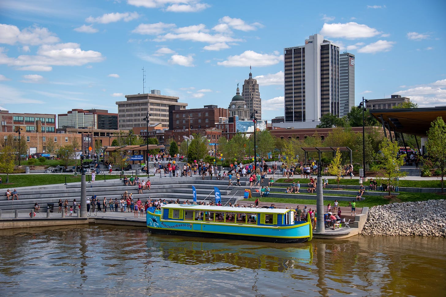 Boat moored at dock in front of public park with city skyline in the background.