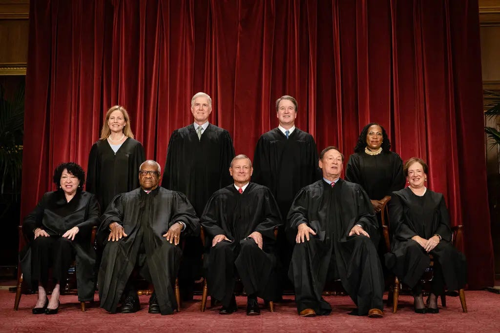 The justices of the Supreme Court posing for a portrait. They are wearing black robes in front of a red curtain.