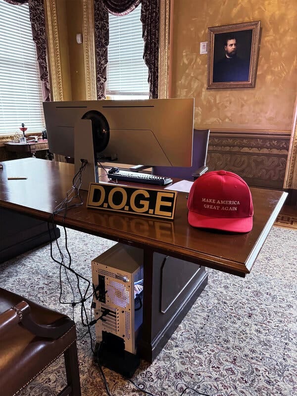 A wooden desk with a computer, a sign reading “D.O.G.E.” and a read “Make America Great” hat.