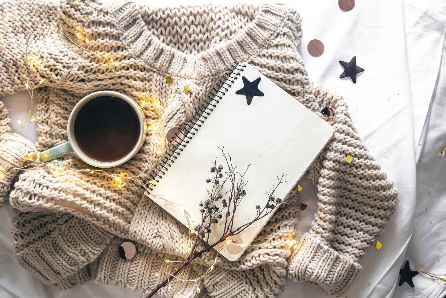 Cozy composition with a notebook, a knitted sweater, fairy lights and a cup of coffee.