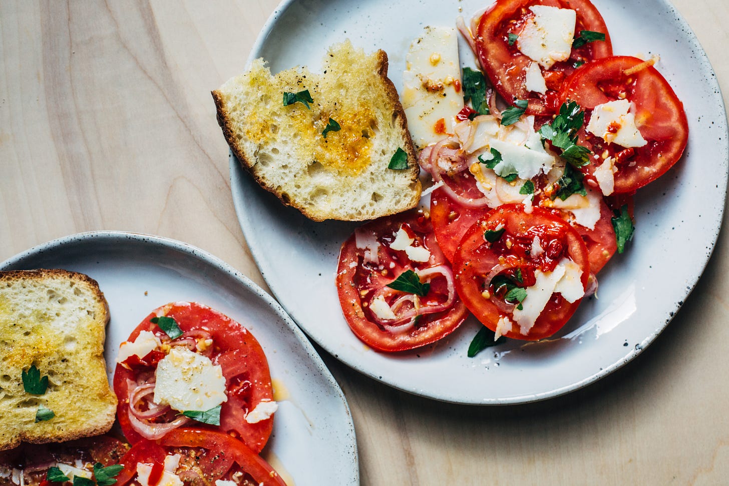 Two plates with tomato salad. The salad is topped with crumbled cheese and herbs, with a slice of bread on the side. 