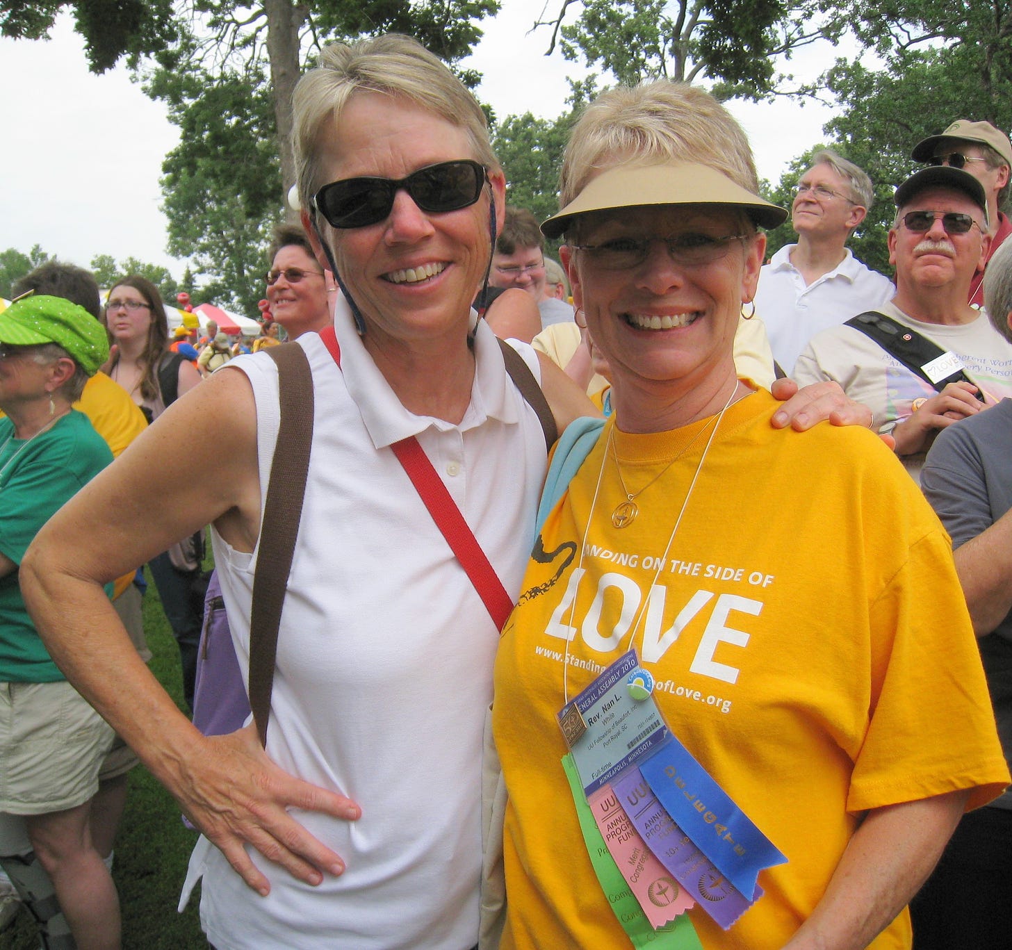 Two women with short-cropped hair--one in a white shirt with one hand on her hip and the other around the other woman. The other wearing a visor and a goldenrod Standing on the Side of Love shirt. 