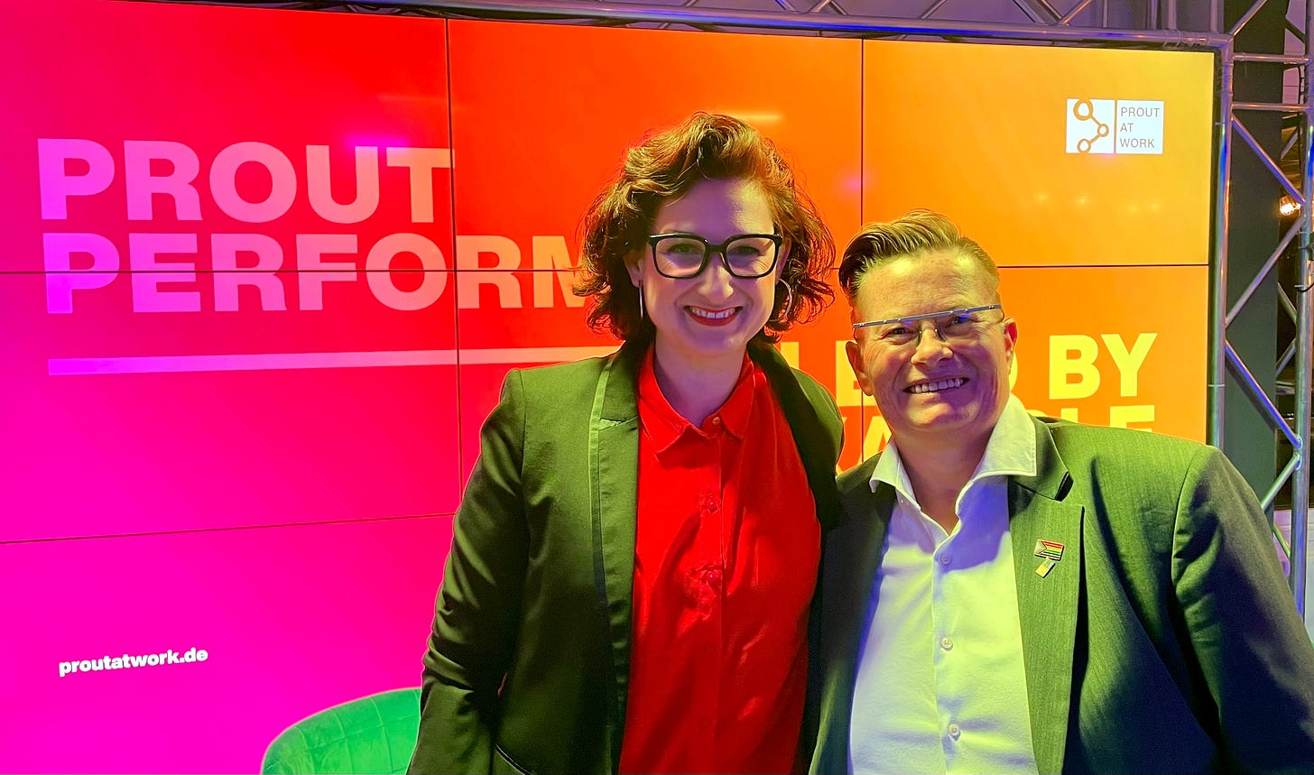 Two individuals stand smiling in front of a vibrant backdrop at the 2022 Prout Performer Networking Event, hosted by the Prout at Work Stiftung. On the left is Ferda Ataman, Germany's Federal Anti-Discrimination Commissioner, advocating for equality and combating discrimination nationwide. On the right, Jay Siegmann, a Prout Performer Top 10 in 2022 and Top 5 in 2021, celebrates their contributions to LGBTQIA+ visibility and workplace inclusion.