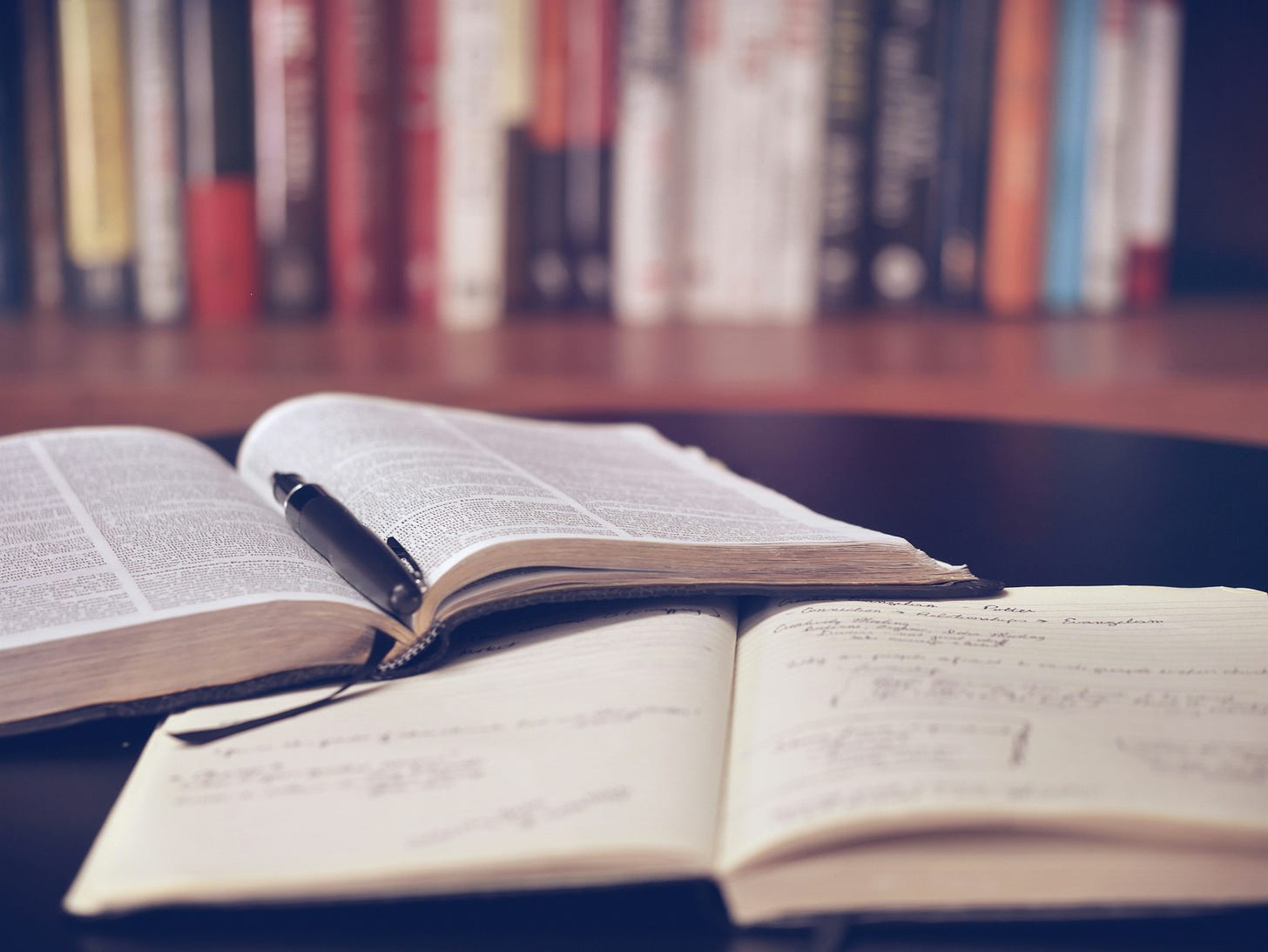 Image of a book, journal, and pen on a desk and books on a shelf behind.