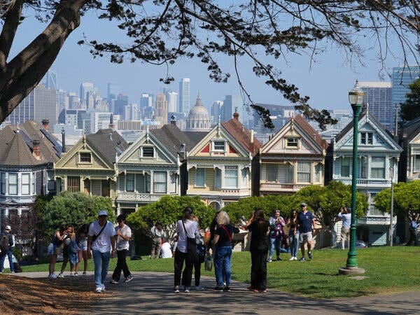On a sunny day with light haze, a couple dozen people mill about a park with the famed Victorian “Painted Ladies” homes nearby and the skyline in the distance.
